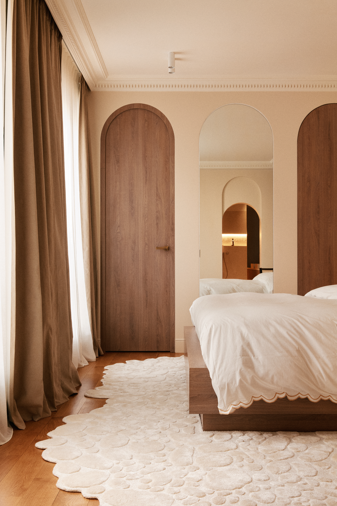 Chambre moderne avec lit blanc, tapis blanc, rideaux beiges, murs beiges, portes en bois arrondies et miroir au mur.