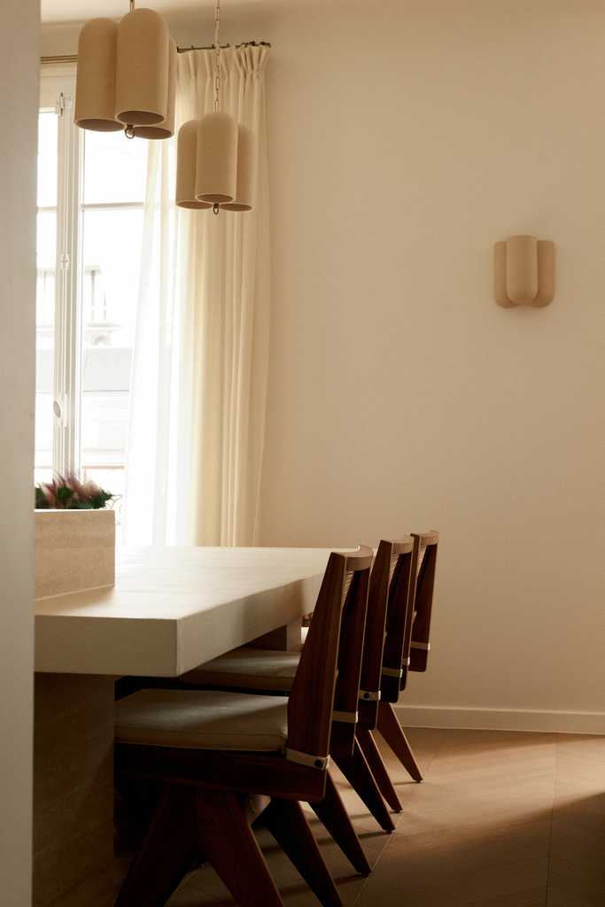 Une table de salle à manger en bois avec des chaises en bois brut, une fenêtre avec des rideaux clairs, un luminaire suspendu et un éclairage mural dans une pièce lumineuse.