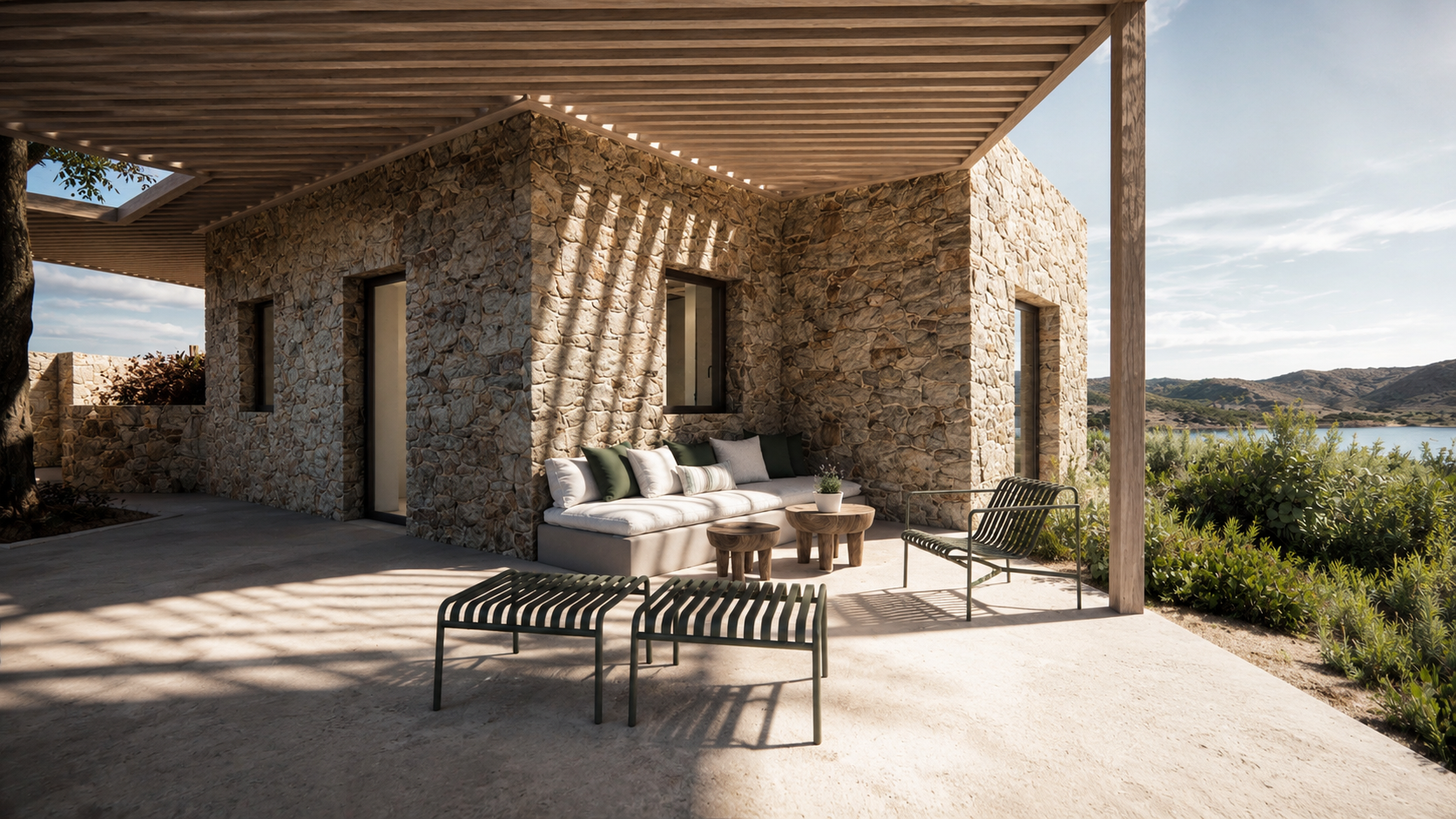 A modern outdoor patio with a stone house, cushioned seating, a small round table, and outdoor chairs, overlooking a lake and mountains in the distance under a partly cloudy sky.