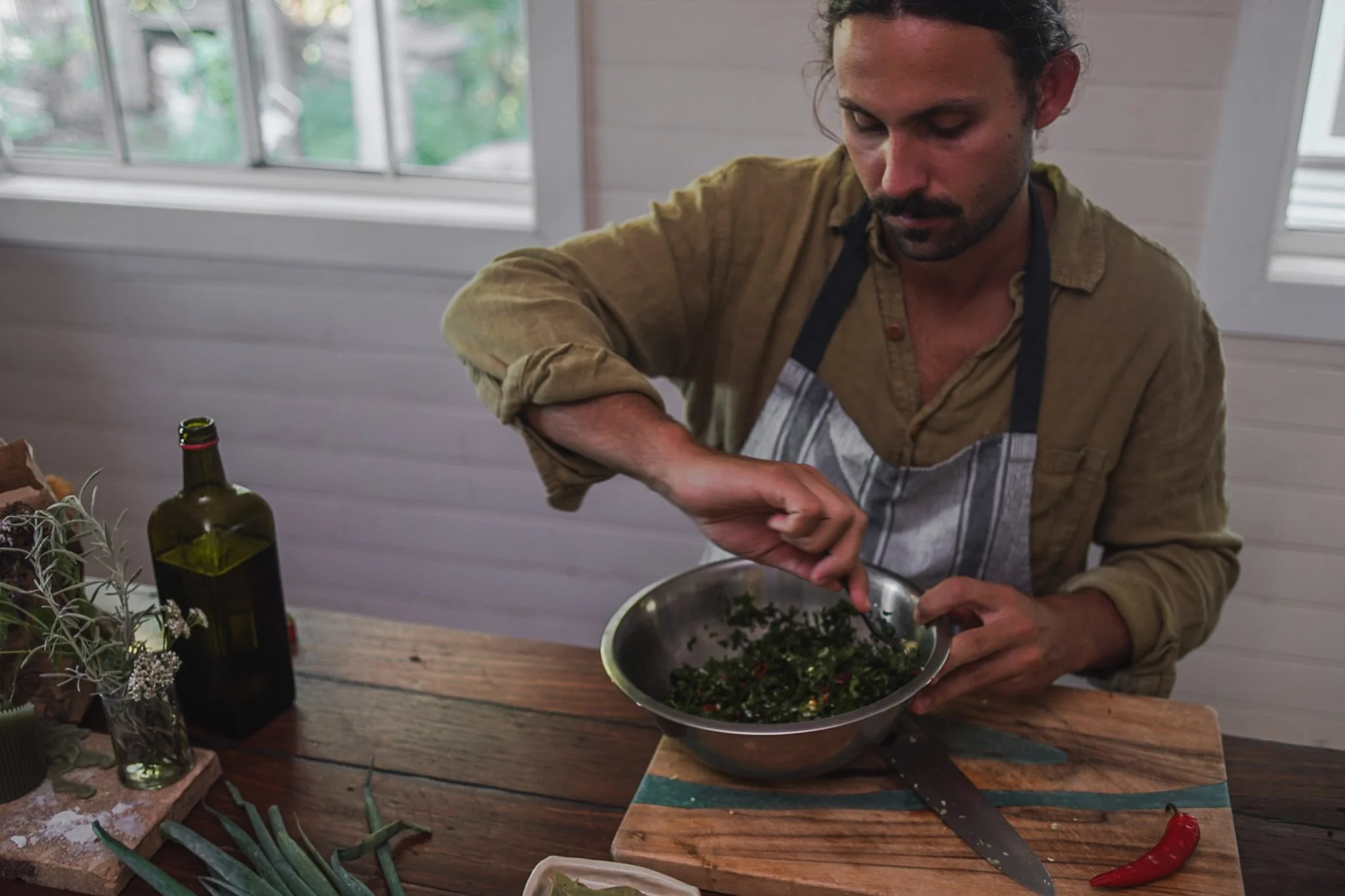 A man wearing a brown shirt and apron preparing food in a bowl on a wooden table near a window, with herbs, a knife, and a bottle of olive oil nearby.