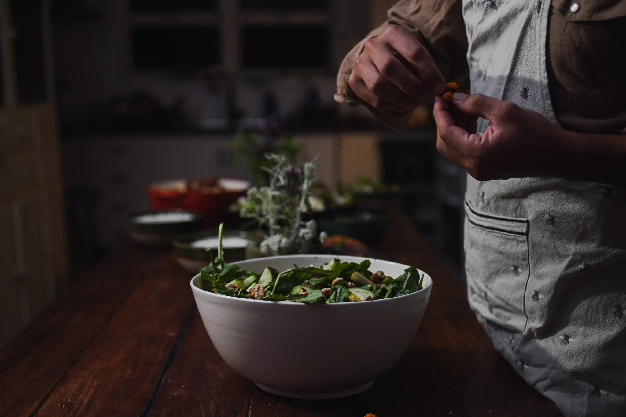 Person preparing a salad in a white bowl with fresh greens and vegetables, in a rustic kitchen setting.
