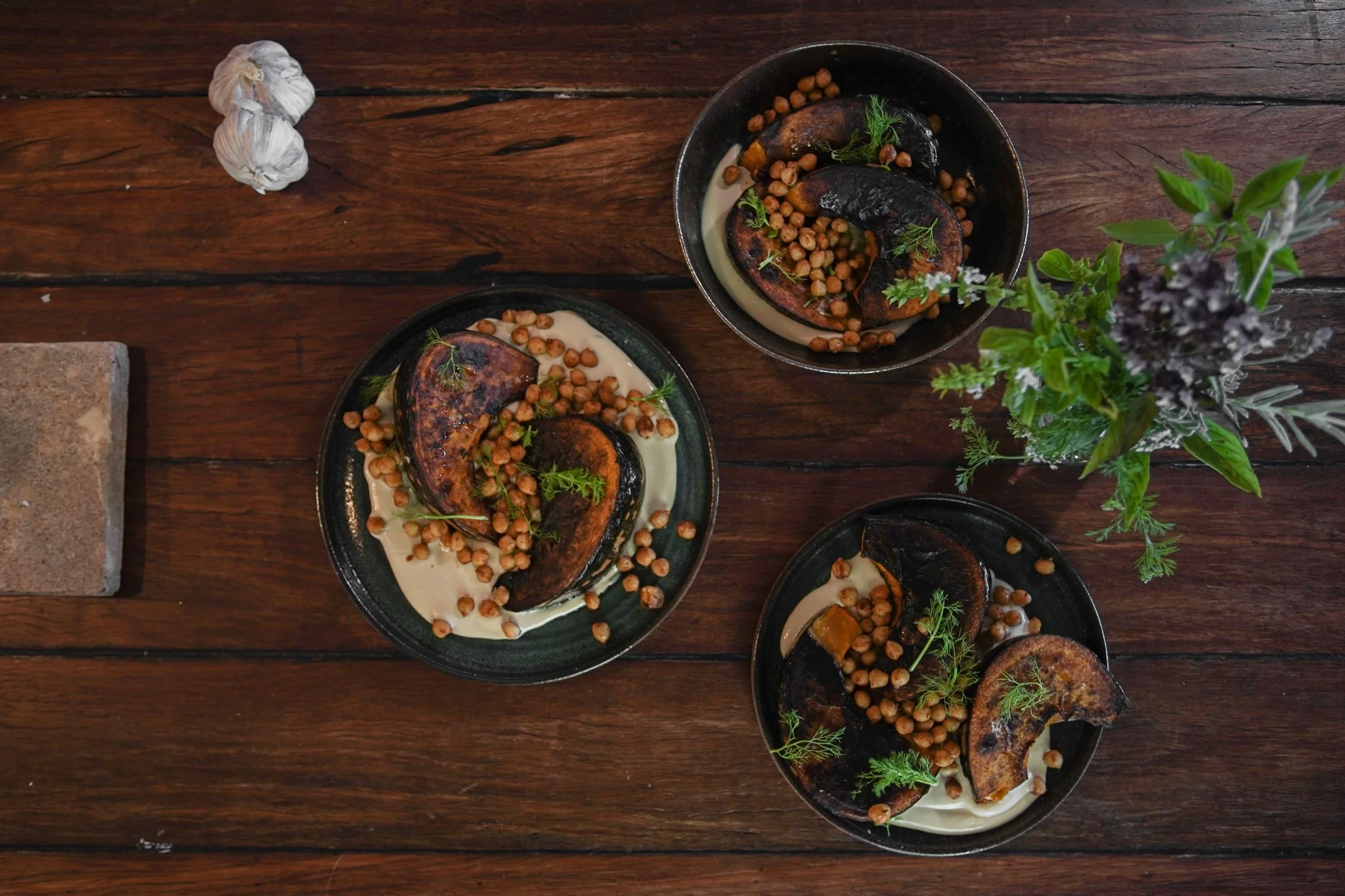 Top view of baked eggplant slices topped with chickpeas, garnished with herbs, served on black plates with creamy sauce, placed on a rustic wooden table with some garlic, a brick, and green plants nearby.