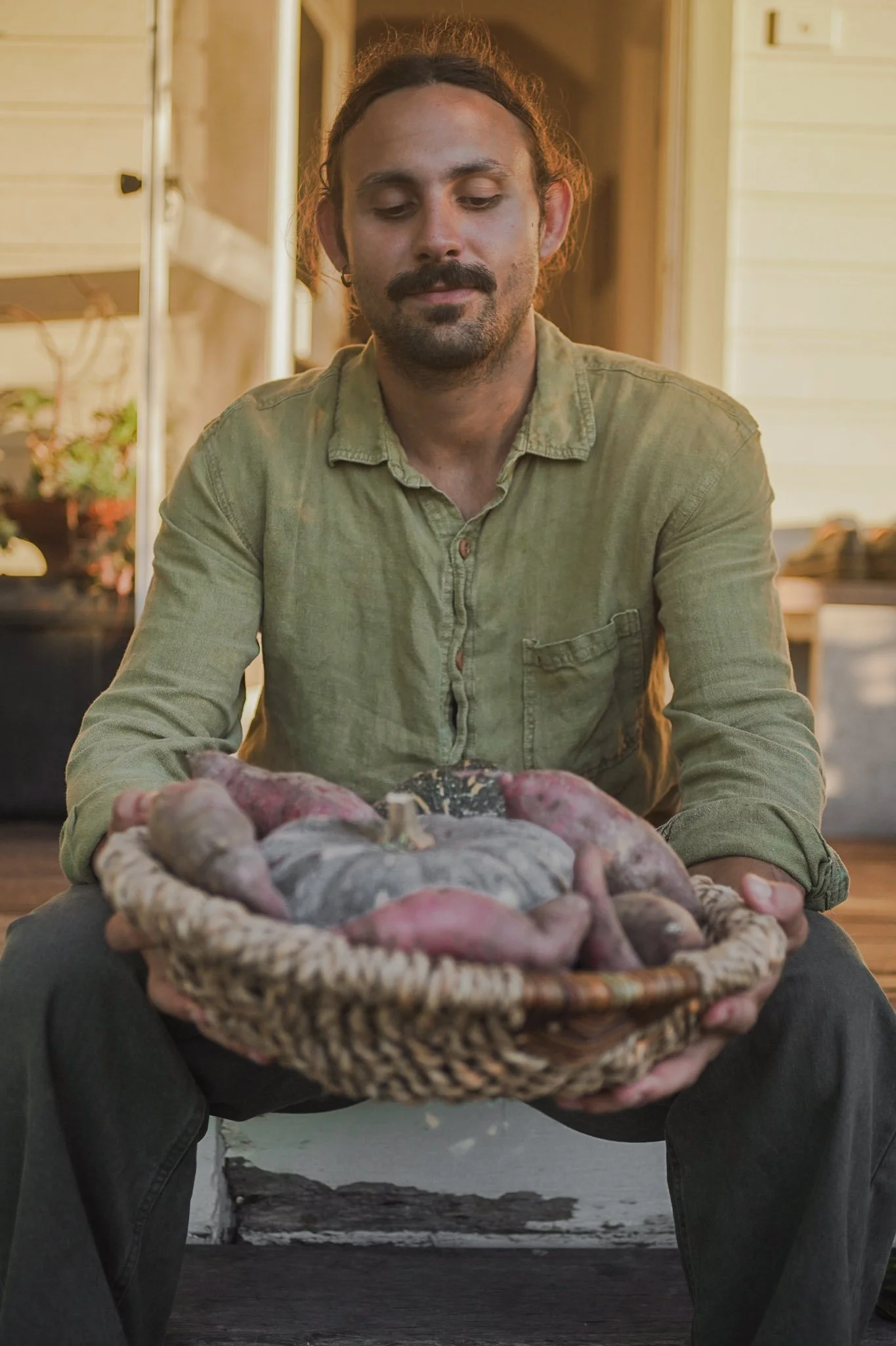 A man with long hair, a mustache, and a beard, sitting in a rustic kitchen, holding a large woven basket filled with freshly harvested sweet potatoes.