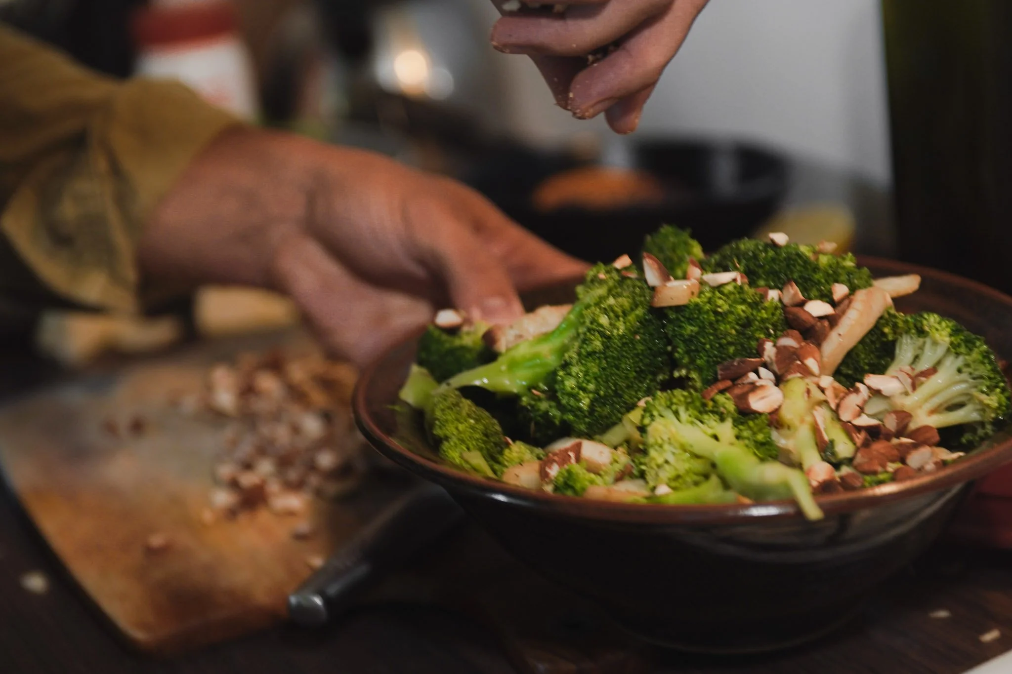 A person's hand sprinkles sliced almonds onto a plate of broccoli and other vegetables at a buffet or food station.