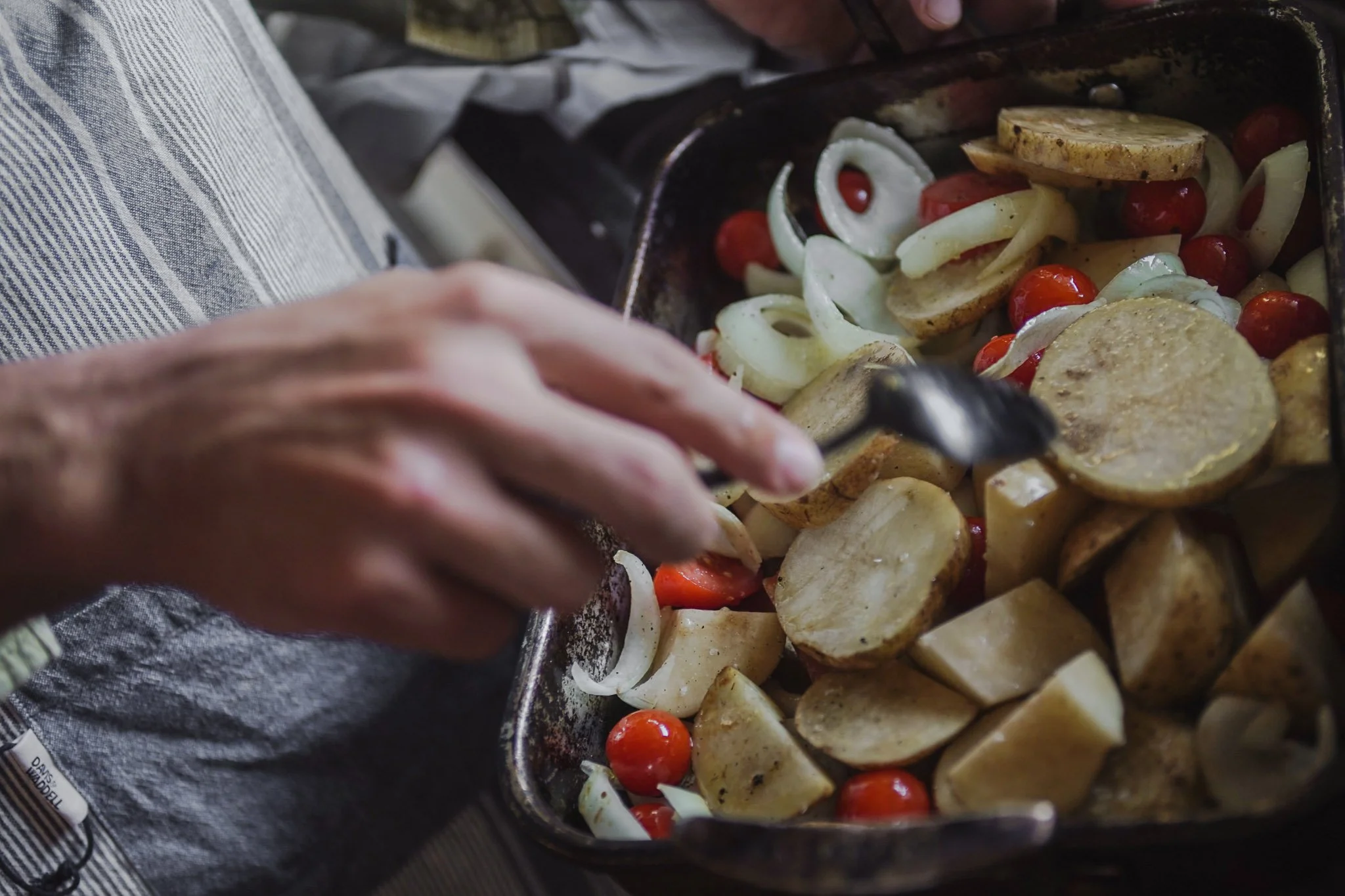 Person cooking vegetables in a cast iron skillet, including cherry tomatoes, onions, and sliced potatoes.