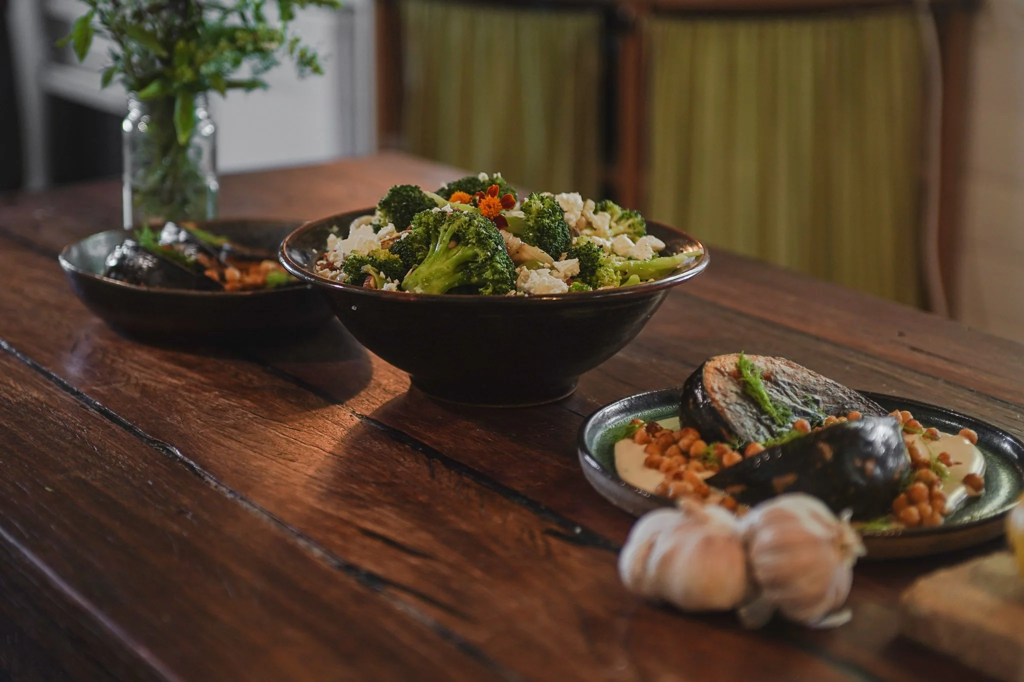 A black bowl filled with broccoli, cauliflower, and decorative flowers on a wooden table. Plates with grilled vegetables and other dishes are partially visible around the bowl.