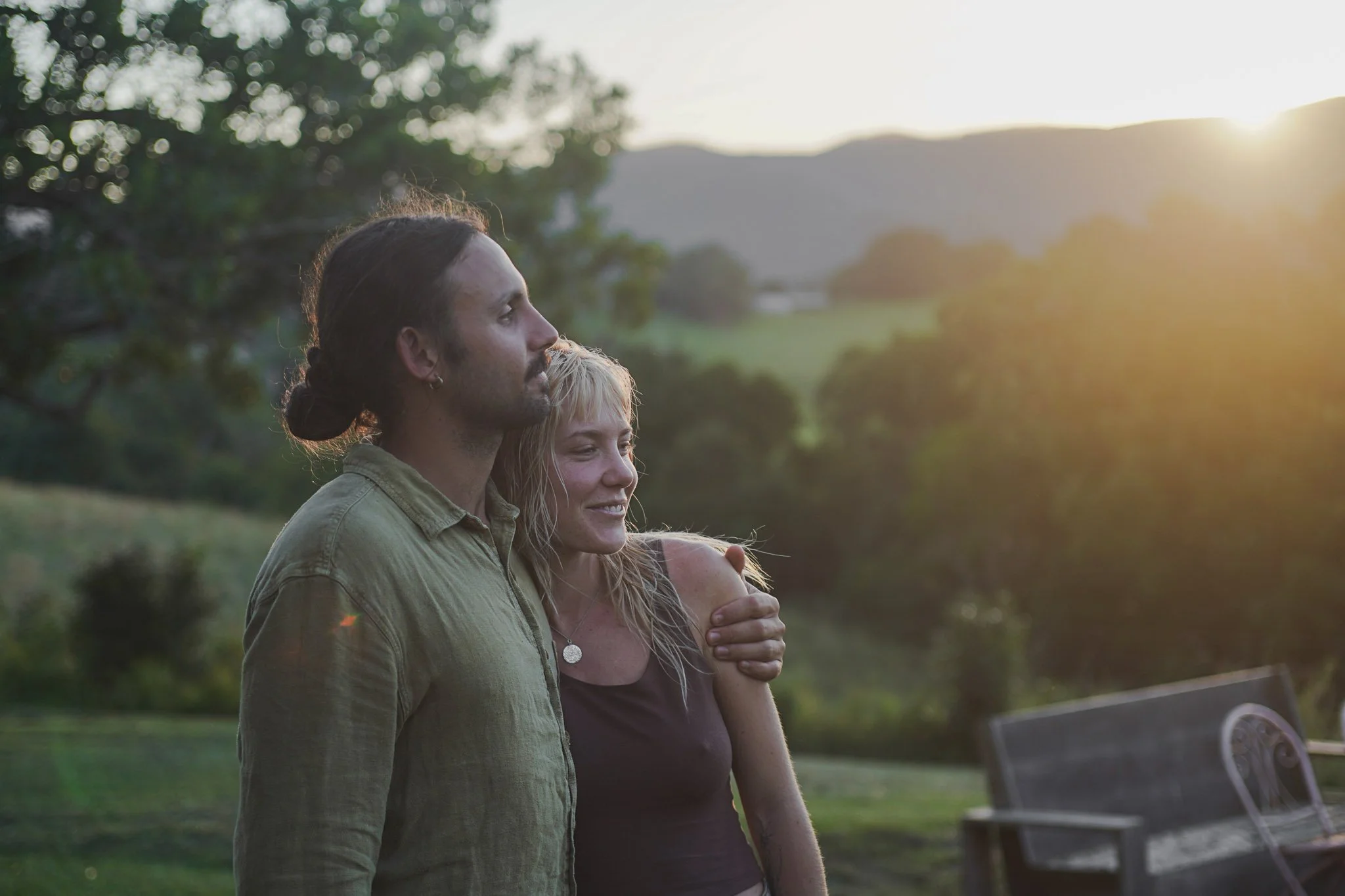 A couple standing outdoors during sunset, embracing each other and looking into the distance, with trees and a park bench in the background.