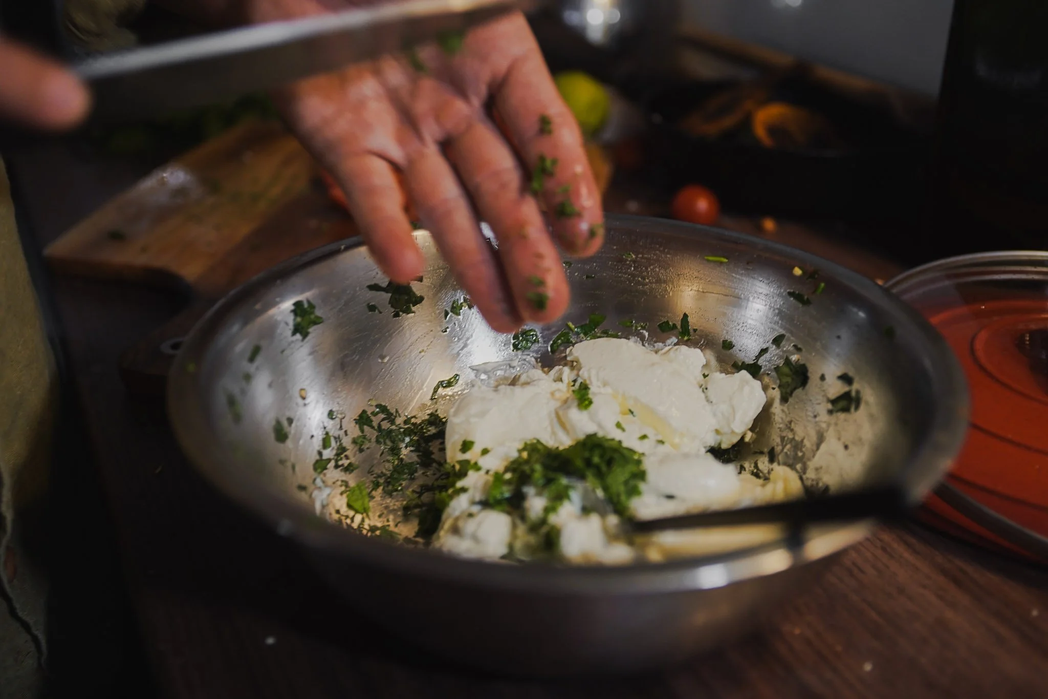 A person's hand is sprinkling chopped herbs over a bowl of cream cheese with herbs, on a wooden countertop.