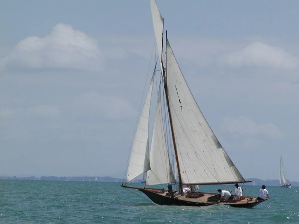 A sailboat with multiple white sails on a body of water, with three people on board and a faint shoreline in the distance.