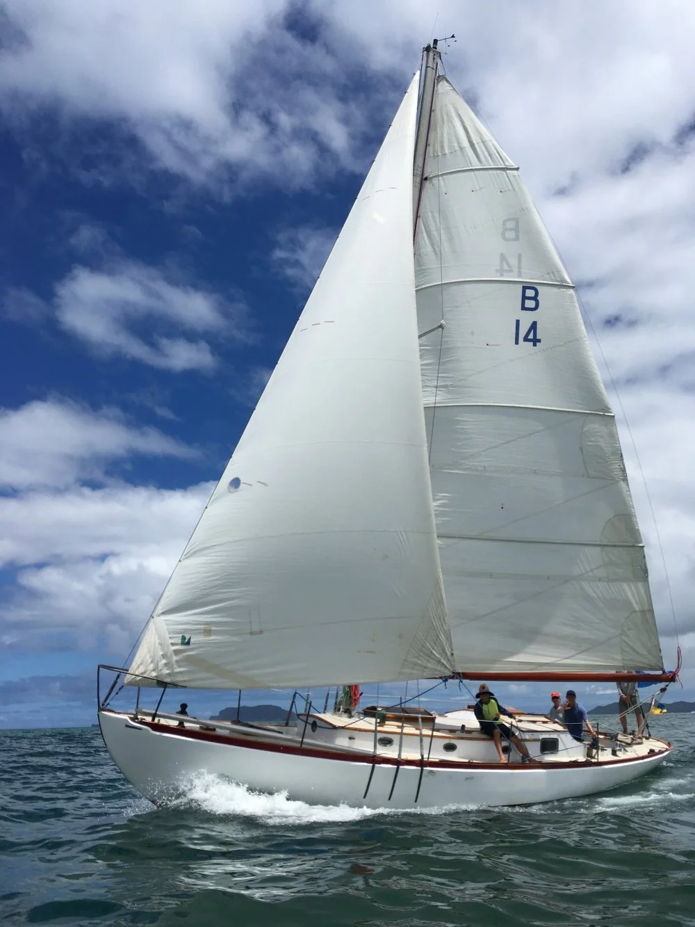 A sailboat with its sails unfurled on a blue ocean under a partly cloudy sky, with a few people on board.