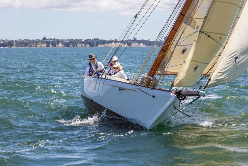 Sailboat with three people sailing on the water, with a distant shoreline in the background.