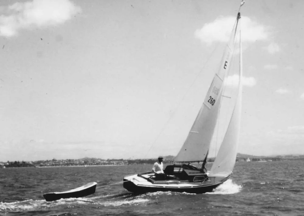 A person sailing a small boat with a large sail on a body of water, pulling a small dinghy behind.