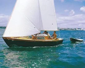 A sailboat with white sails on the ocean, with two people on board and another smaller boat nearby, under a partly cloudy sky.