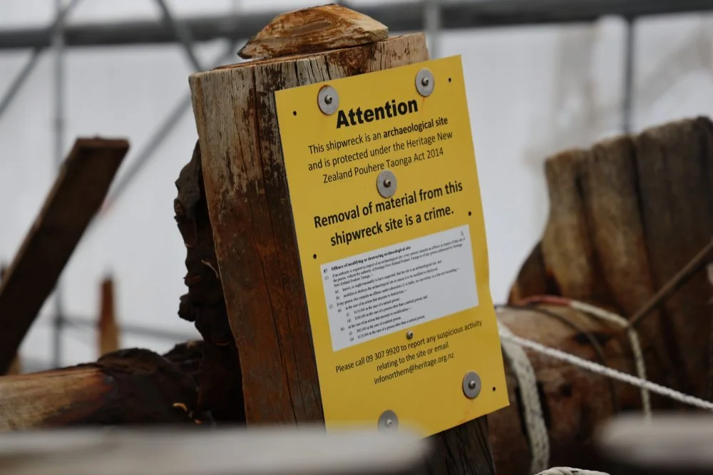 A yellow caution sign attached to a wooden post at an archaeological shipwreck site, warning against removing materials. The sign mentions the site is protected under New Zealand law and encourages reporting suspicious activity.