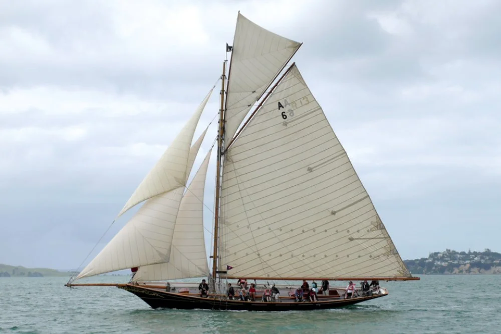 A sailing ship with large white sails on the water during cloudy weather, with land visible in the background.