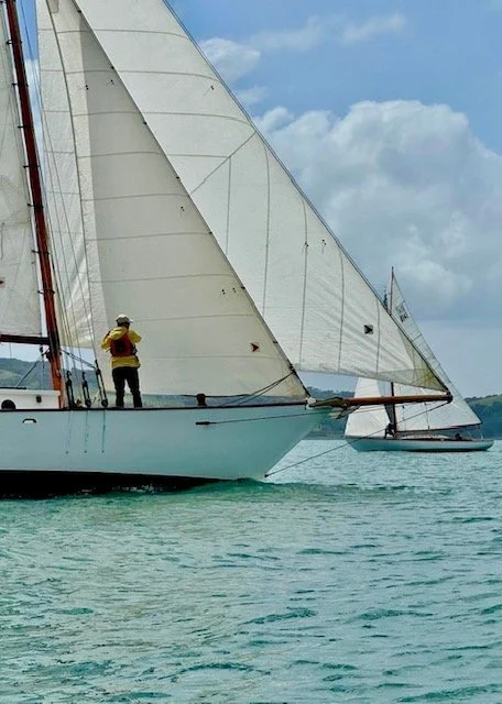 A sailboat with two large sails on the water during the daytime, with a person standing on the deck wearing a hat and life vest.