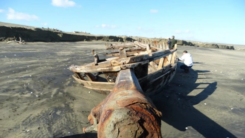 Two people examining the wreckage of an old, weathered wooden boat on a sandy beach under a clear sky.