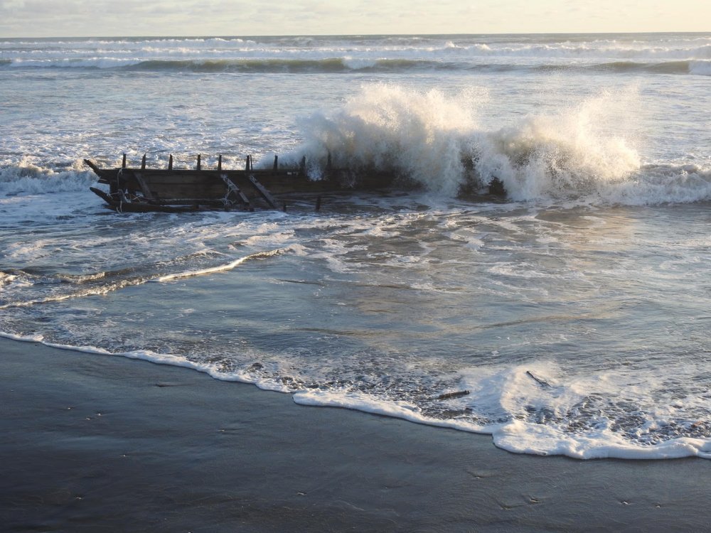 Wreckage of a boat partially submerged as waves crash over it on a sandy beach.