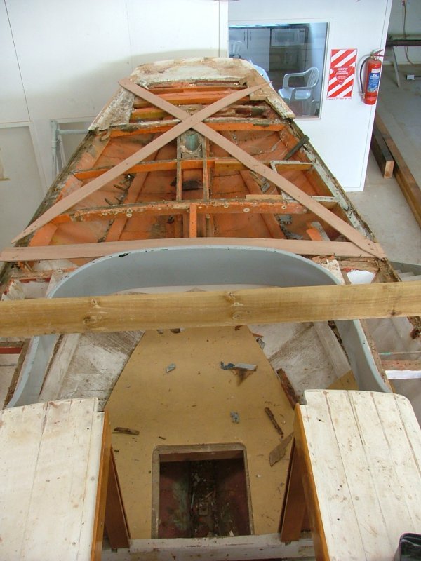 The image shows the interior of a boat under renovation, with exposed wooden framework, support beams, and construction materials inside a workshop.