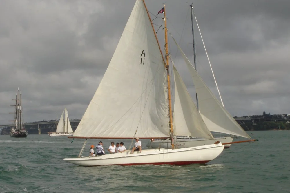 Sailing boat with white sails on ocean with cloudy sky, multiple people on board, in front of a bridge and other sailboats.