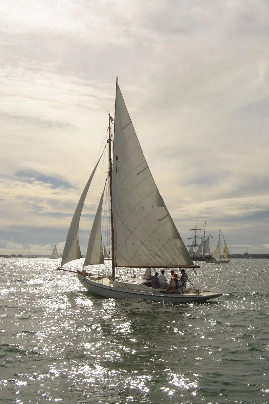 Sailboat with people on board sailing on water with other sailboats and ships in the background under a cloudy sky.