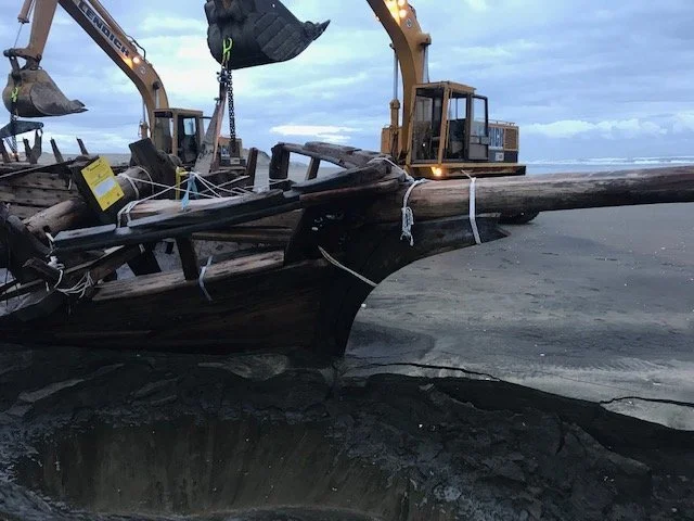 Large hole in the ground with a wooden boat resting on the edge, surrounded by excavators at a construction site.