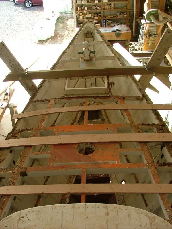 View of a boat hull inside a workshop, with wooden frames and tools around, looking down its length.