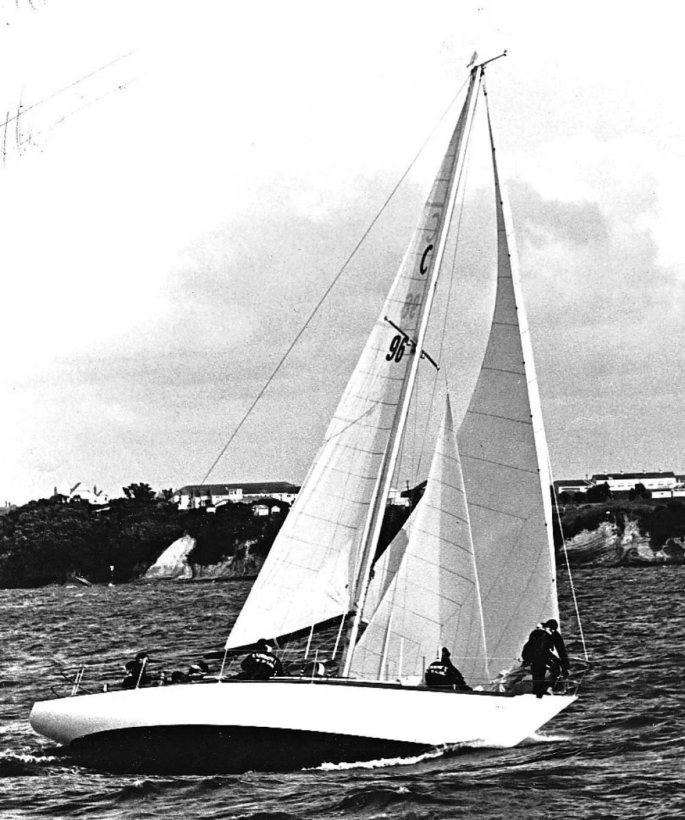 Black and white photo of a sailboat on the water with four crew members. The boat has large sails and a coastline with houses in the background.