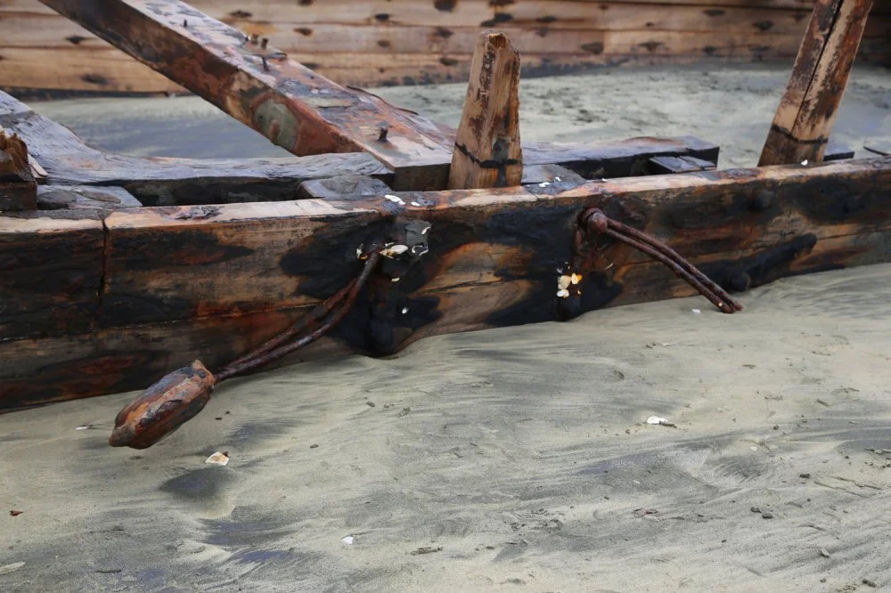 Close-up of a weathered, broken wooden boat resting on sandy ground, with some metal wires attached, indicating it is no longer in use.