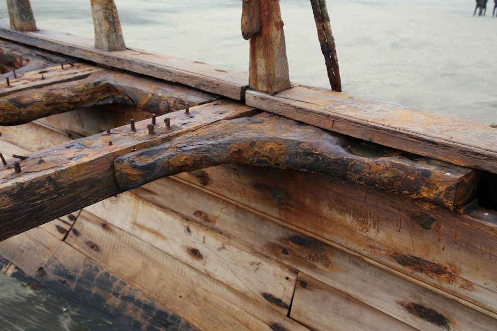 Close-up of a weathered, rusty wooden boat under construction or repair, with nails and planks visible, on water.