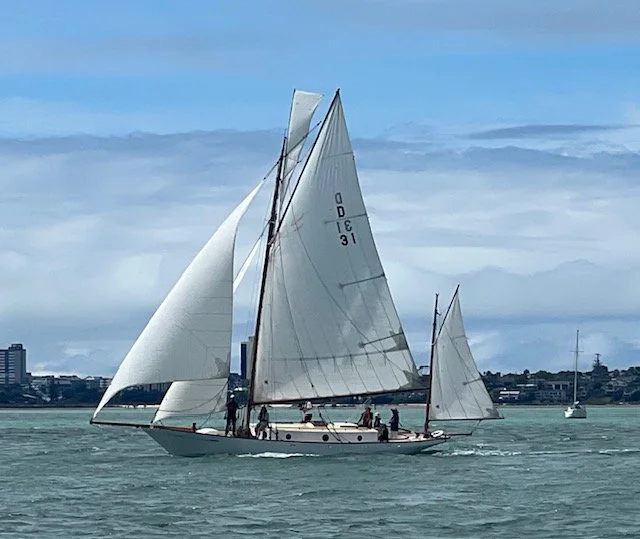 A large sailboat with multiple white sails on the water, with a city skyline in the background and cloudy sky above.
