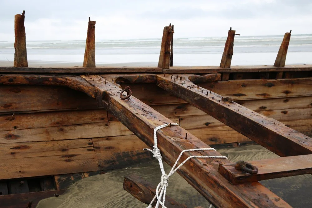 Old wooden boat hull on the beach under a cloudy sky, with protruding ribs and a tied rope.