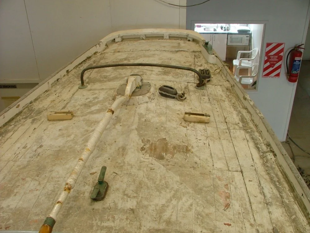 Close-up of the top of a boat hull that is weathered and dirty, with tools and equipment resting on it, and a fire extinguisher and safety signs on the wall nearby.