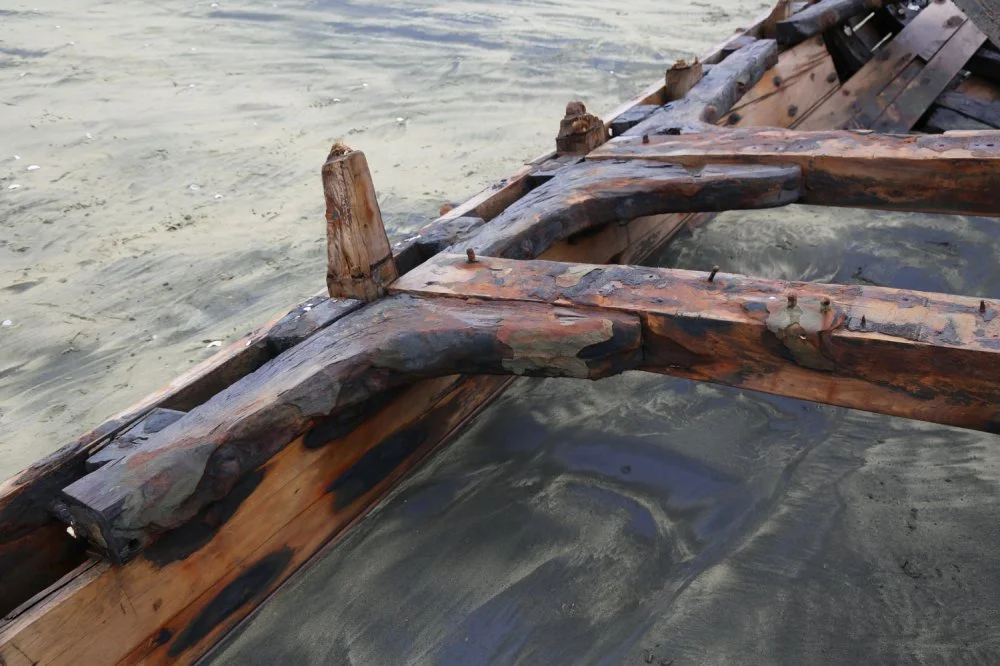 Close-up of a damaged wooden boat on a muddy shoreline, showing broken and burnt wooden planks.