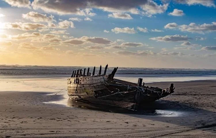 An old, abandoned wooden shipwreck, The Daring, on a sandy beach during sunset with a partly cloudy sky.