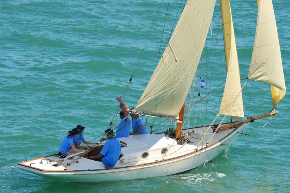 A group of five people sailing on a sailboat in the water, with some people sitting and others standing and waving.
