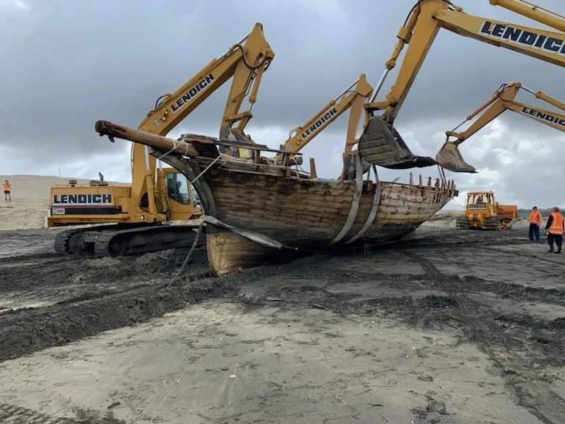 The excavators are lifting the boat and walking up the beach while the bulldozer assists in pulling.