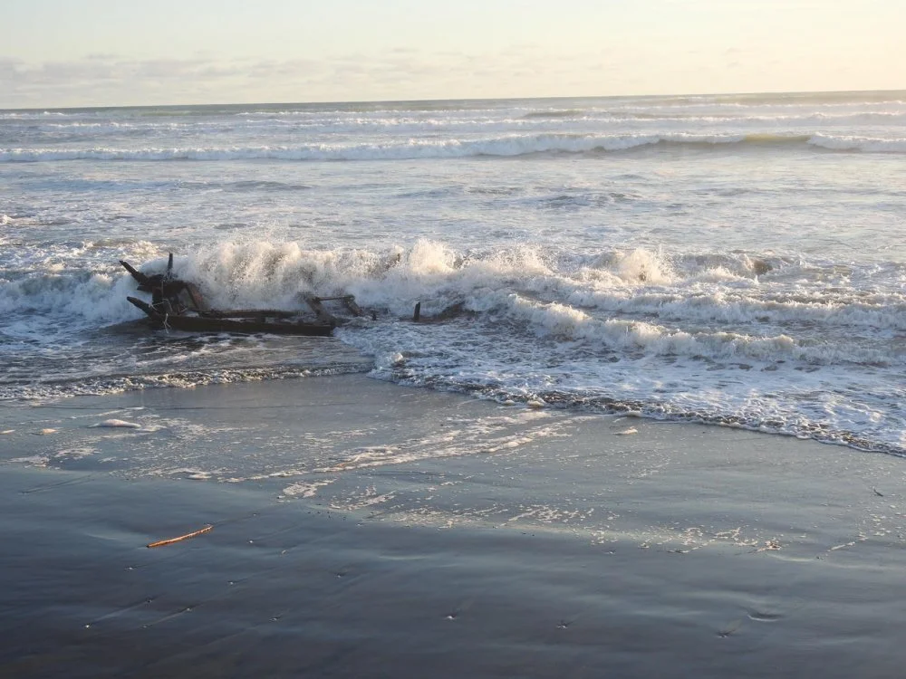 Wreckage of a small boat washed up on wet sandy beach with waves crashing around it, overcast sky.