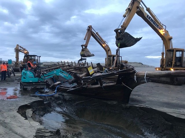 Multiple excavators working on a sunken boat on a muddy shoreline during daytime