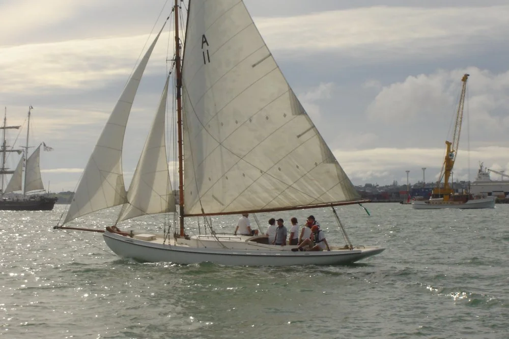A sailboat with six people onboard sailing on the water, with other sailboats and ships in the background during cloudy weather.