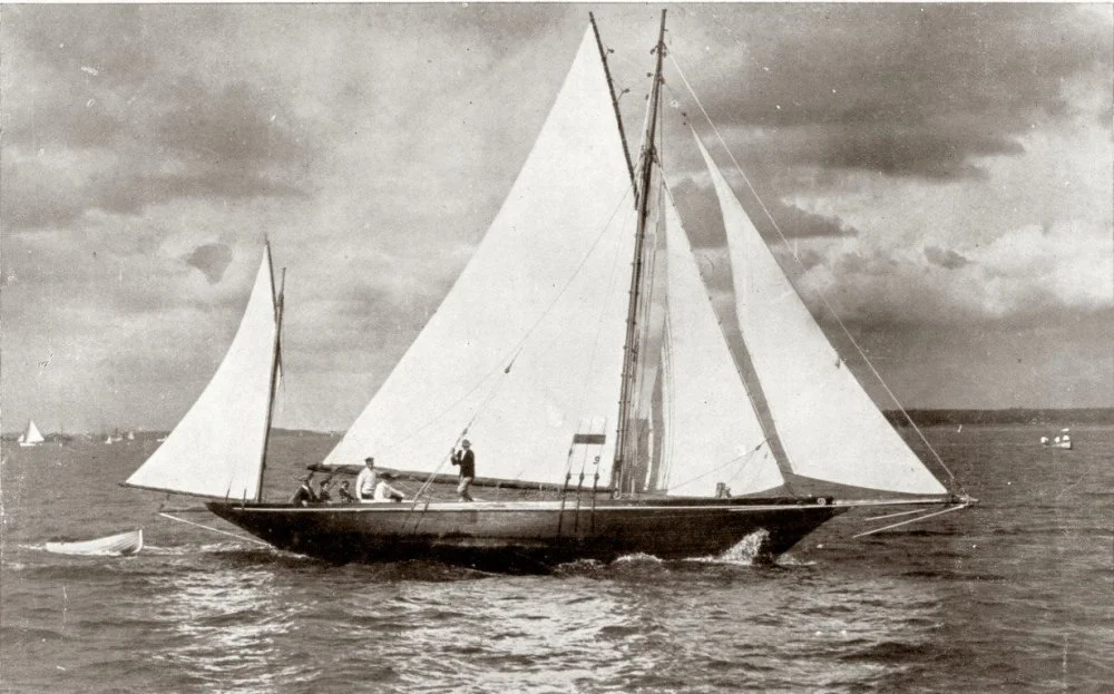 A vintage black-and-white photo of a sailboat with large sails on the water, with some people on board and a small rowboat nearby.