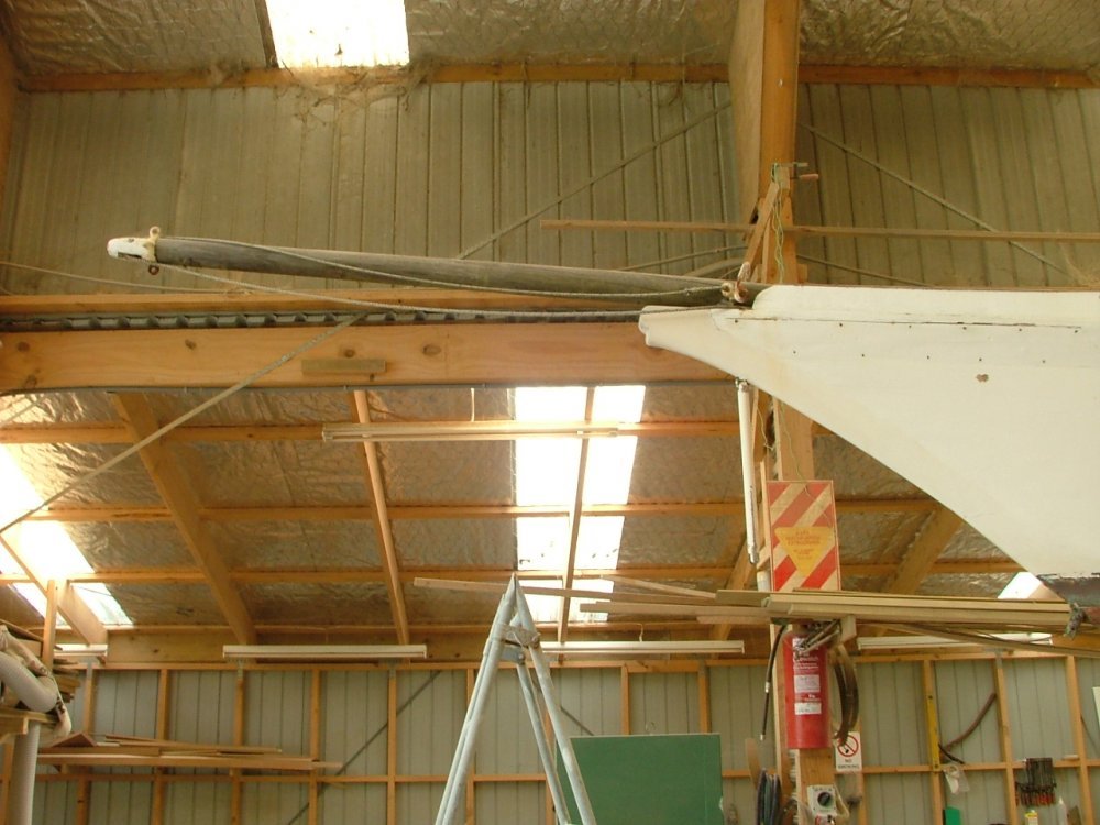 The interior of a warehouse or workshop with a high ceiling, wooden beams, insulation, and fluorescent lights. There is a tall metal ladder, a fire extinguisher, and various construction or industrial equipment.