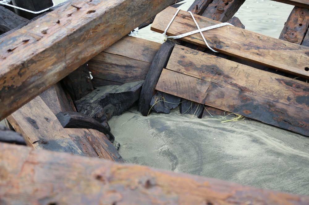 Close-up of a damaged wooden boat on a sandy beach, showing broken and weathered planks and beams.
