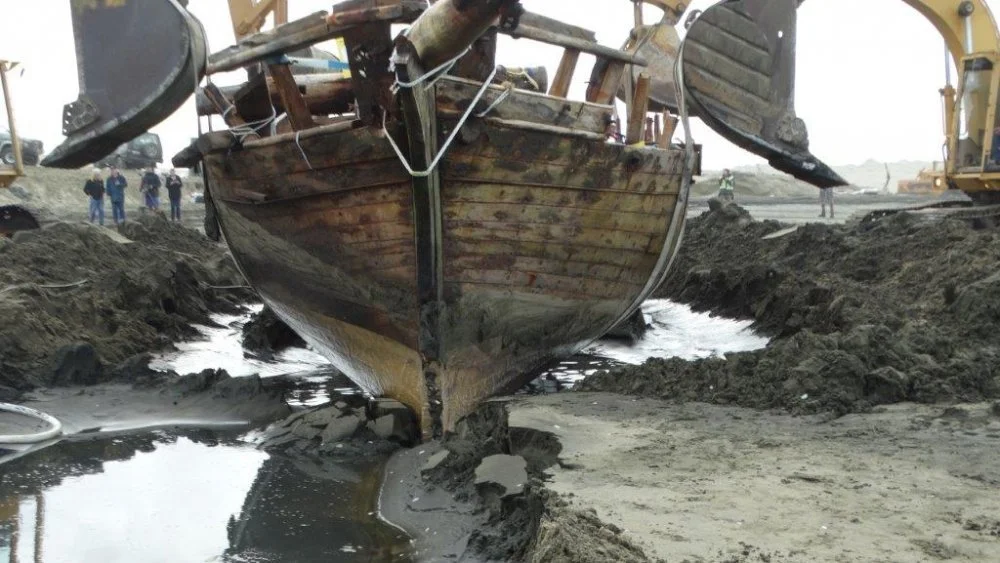 A wooden boat is out of the water on a construction site with bulldozers and workers in the background.