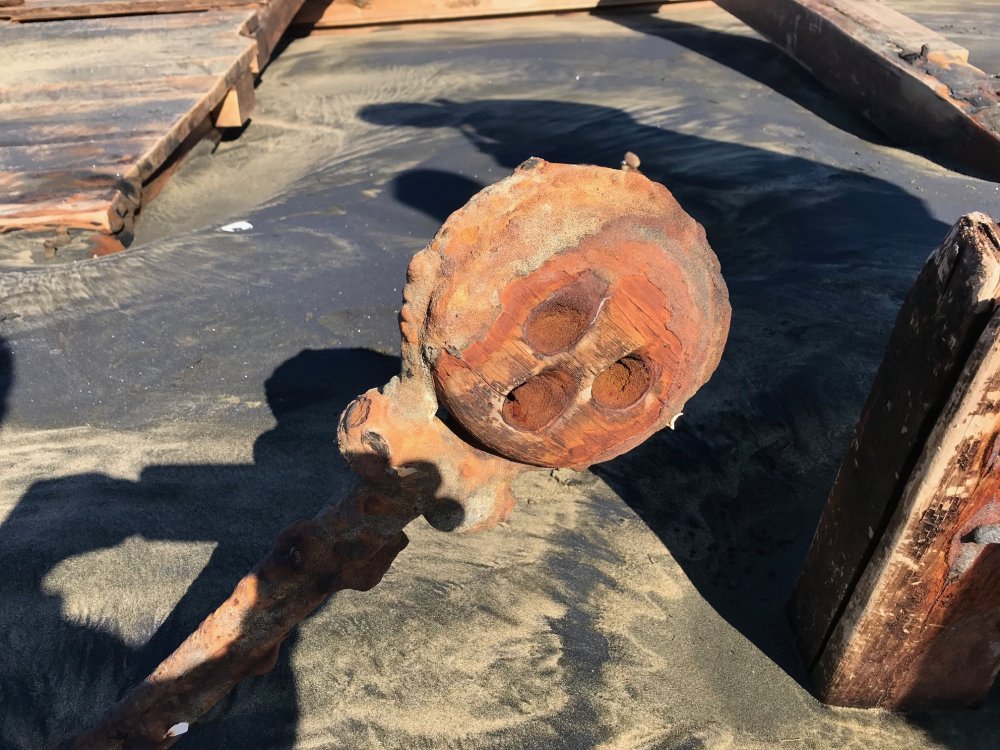 Close-up of a rusted, old electrical outlet with three circular holes, mounted on a rusty metal pipe, against a sandy surface with wooden planks in the background.