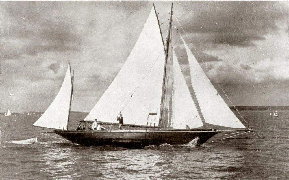 A black and white photo of a sailboat with sails unfurled, featuring several people on board, sailing on open water with a few other boats in the distance under cloudy skies.