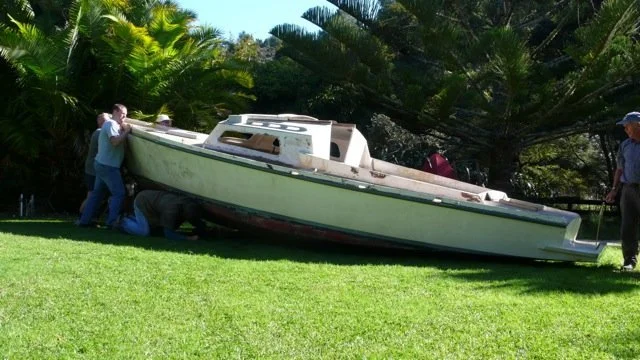 People working on or inspecting a large sailboat or yacht on a grassy area with lush green trees in the background.