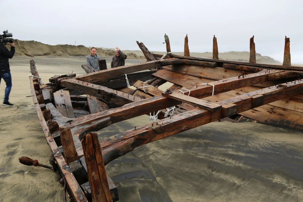 A large wooden shipwreck on a sandy beach with three people and a cameraman nearby, with cliffs in the background.