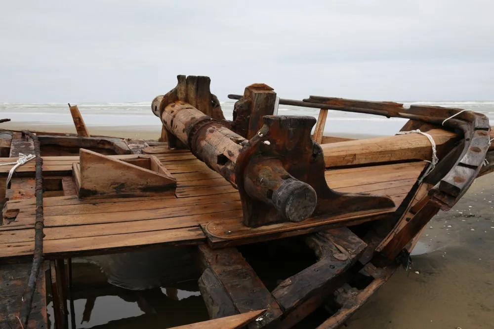 A weathered, old wooden boat on a sandy beach, with parts of the boat appearing damaged or decayed.