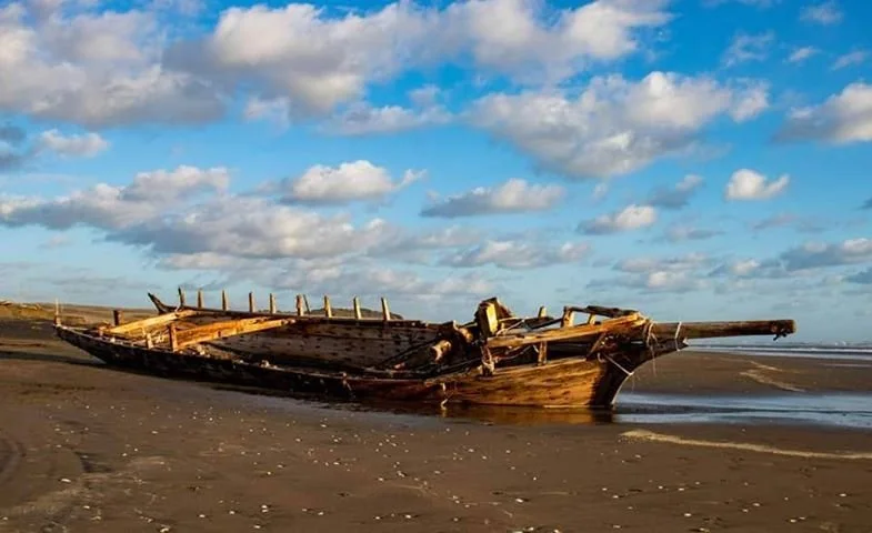 The Daring, wooden shipwreck, on a sandy beach with blue sky and scattered clouds.